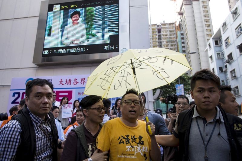 Pro-democracy activist Tam Tak-chi is escorted away by police after confronting government supporters as Chief Secretary Carrie Lam is pictured on a giant television screen during a promotional event she led on electoral reform, in Hong Kong, China on April 25, 2015. (REUTERS File Photo)