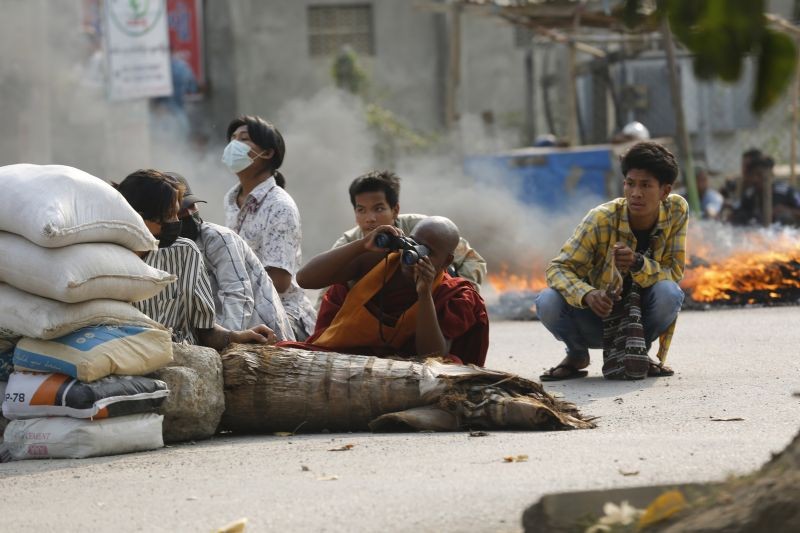 A Buddhist monk uses binoculars while with other men squatting behind a road barricade on March 22, 2021, in Mandalay, Myanmar. (AP/PTI Photo)